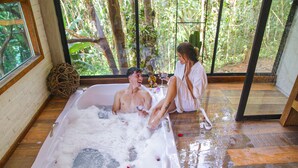 A room with a large bathtub filled with bubbles, a wooden floor, and a view of a forest through large windows. - Pousada irreplaceable (Bocaina de Minas)