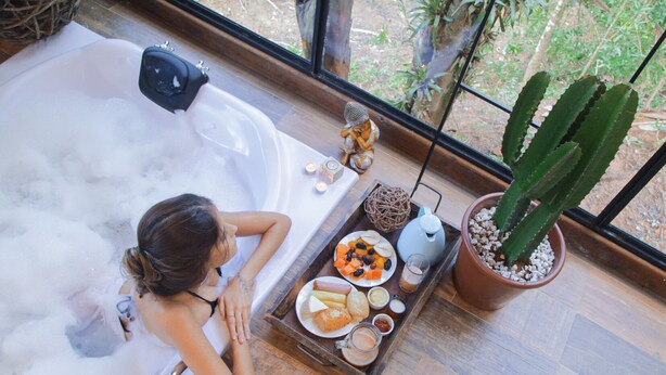 A woman relaxing in a bathtub with a tray of food and drinks beside her. - Pousada irreplaceable (Bocaina de Minas)