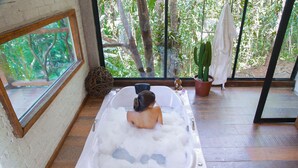 A spa room with a bathtub filled with bubbles, a potted plant, and a window with a view of trees. - Pousada irreplaceable (Bocaina de Minas)