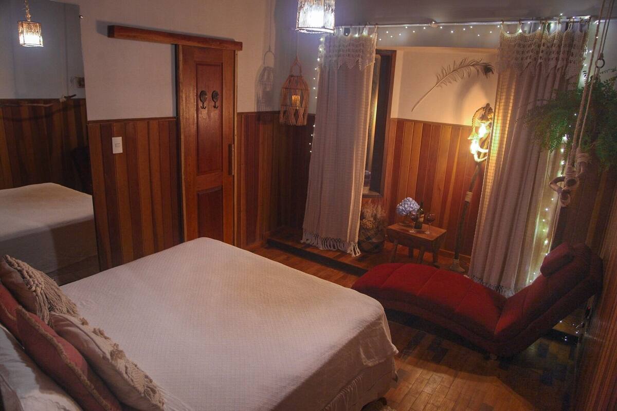 Una habitación con una cama, una silla roja, una mesita con una lámpara y una puerta con marco de madera.