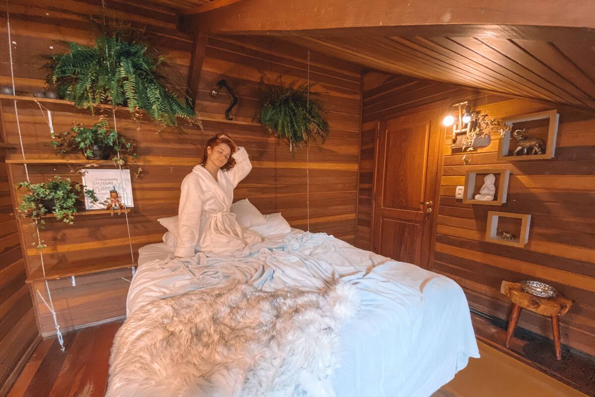 Una habitación acogedora de madera con una cama, un taburete pequeño y estantes con plantas y cuadros en las paredes.
