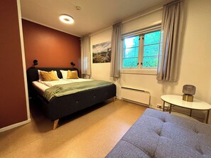 A bedroom with a bed, a radiator, a window with curtains, and a small round table.
