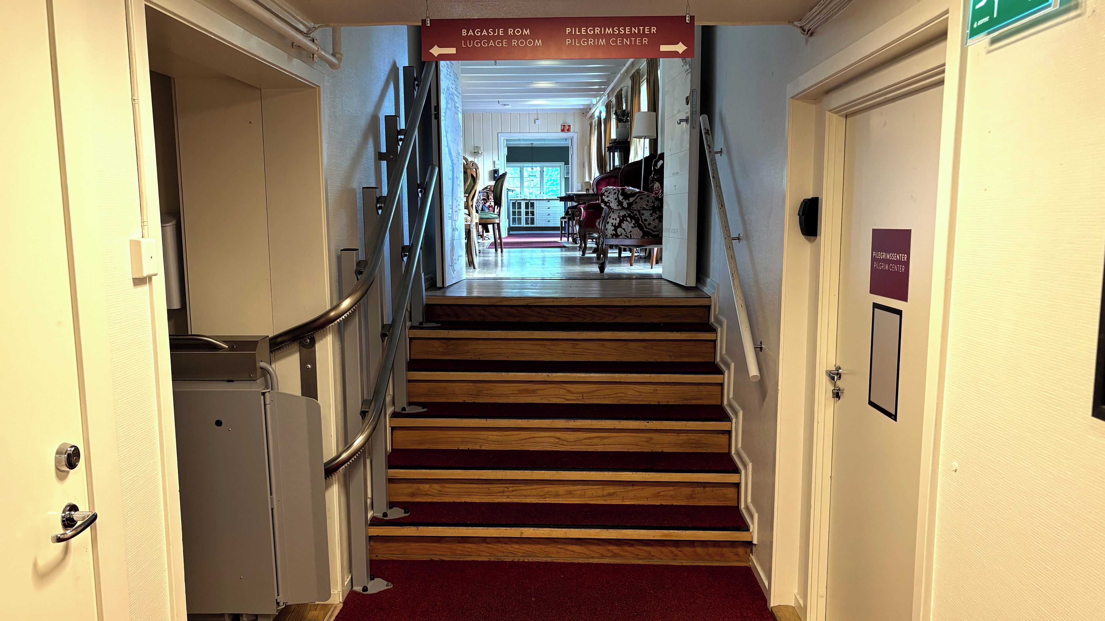 A hotel hallway with a red carpet, wooden stairs, and signs indicating the luggage room and pilgrim center.