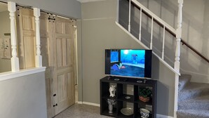 A living room with a television on a stand, a staircase, and a door with a curtain. - 11-Guests/Airport -Minute Away/Pet-Welcome/4-BDR/2-FBA/5-QBED! (Oklahoma City)