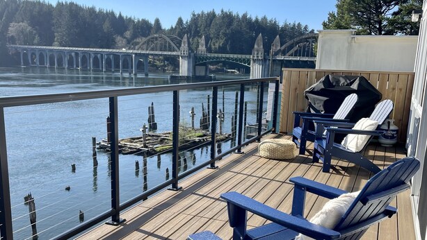 Eine Terrasse mit zwei Adirondack-Stühlen, einem Grill und Blick auf eine Brücke über dem Wasser.