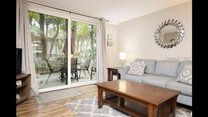 Un salon doté d’une porte-fenêtre coulissante donnant sur une terrasse, d’un canapé gris agrémenté de coussins à motifs, d’une table basse en bois et d’un miroir décoratif accroché au mur.