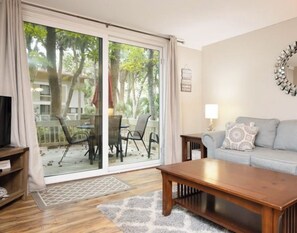 Een woonkamer met een schuifdeur naar een terras, een grijze bank met decoratieve kussens, een houten salontafel en een decoratieve spiegel aan de muur.