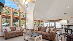 A spacious living room with two sofas, a coffee table, and a desk with a lamp. Large windows provide natural light and views of the outdoors.