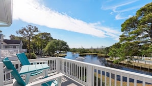 A wooden deck with white railings overlooking a calm river, surrounded by trees and a distant bridge.