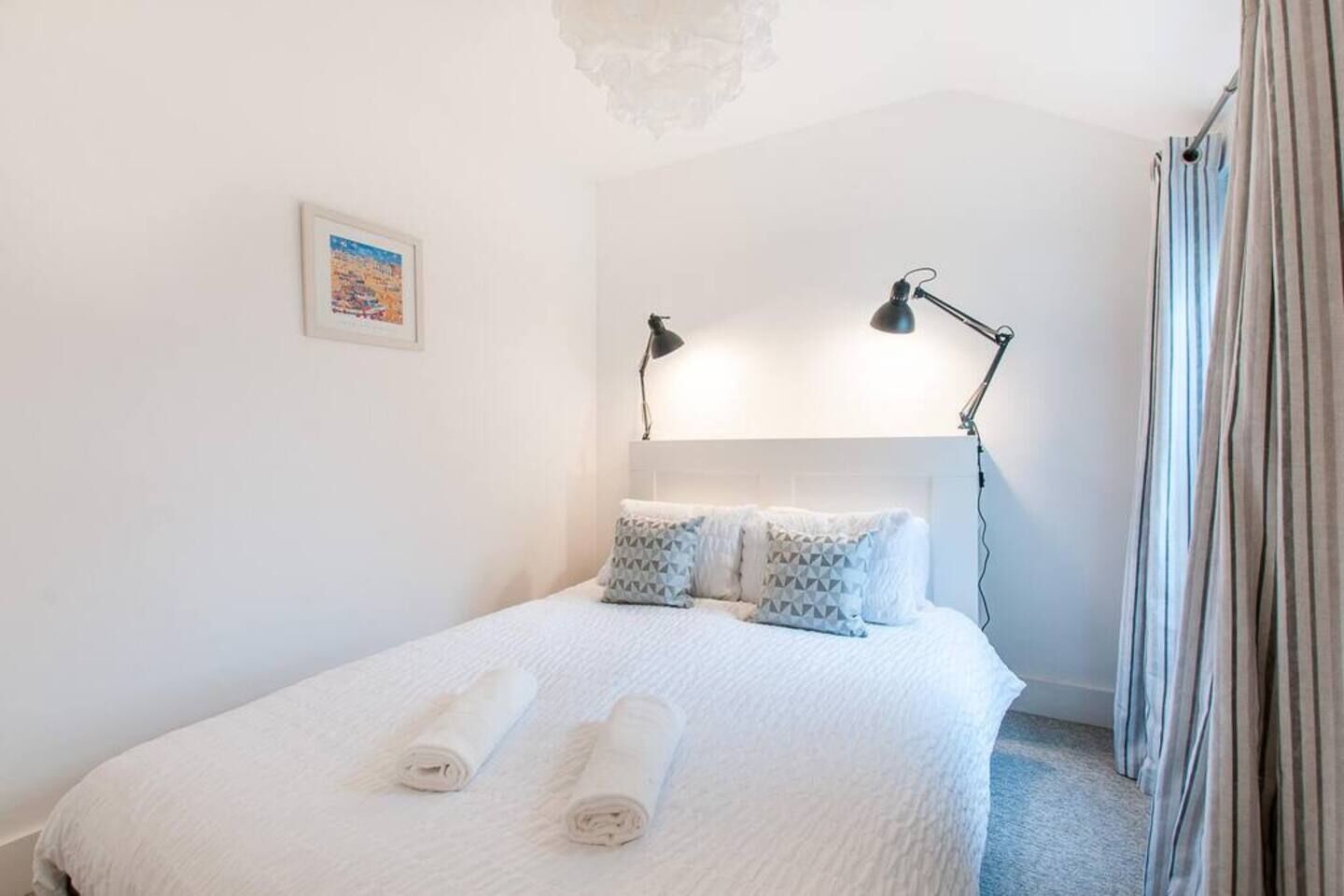 Una cama bien hecha con almohadas y toallas enrolladas, un cabecero, dos lámparas de noche y un cuadro en la pared.