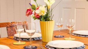 A dining table set with white plates, blue patterned placemats, and clear wine glasses. A yellow vase with flowers is centered on the table. - Coal Miners Cottage - Moments to Seafront & Deal Town (Deal)