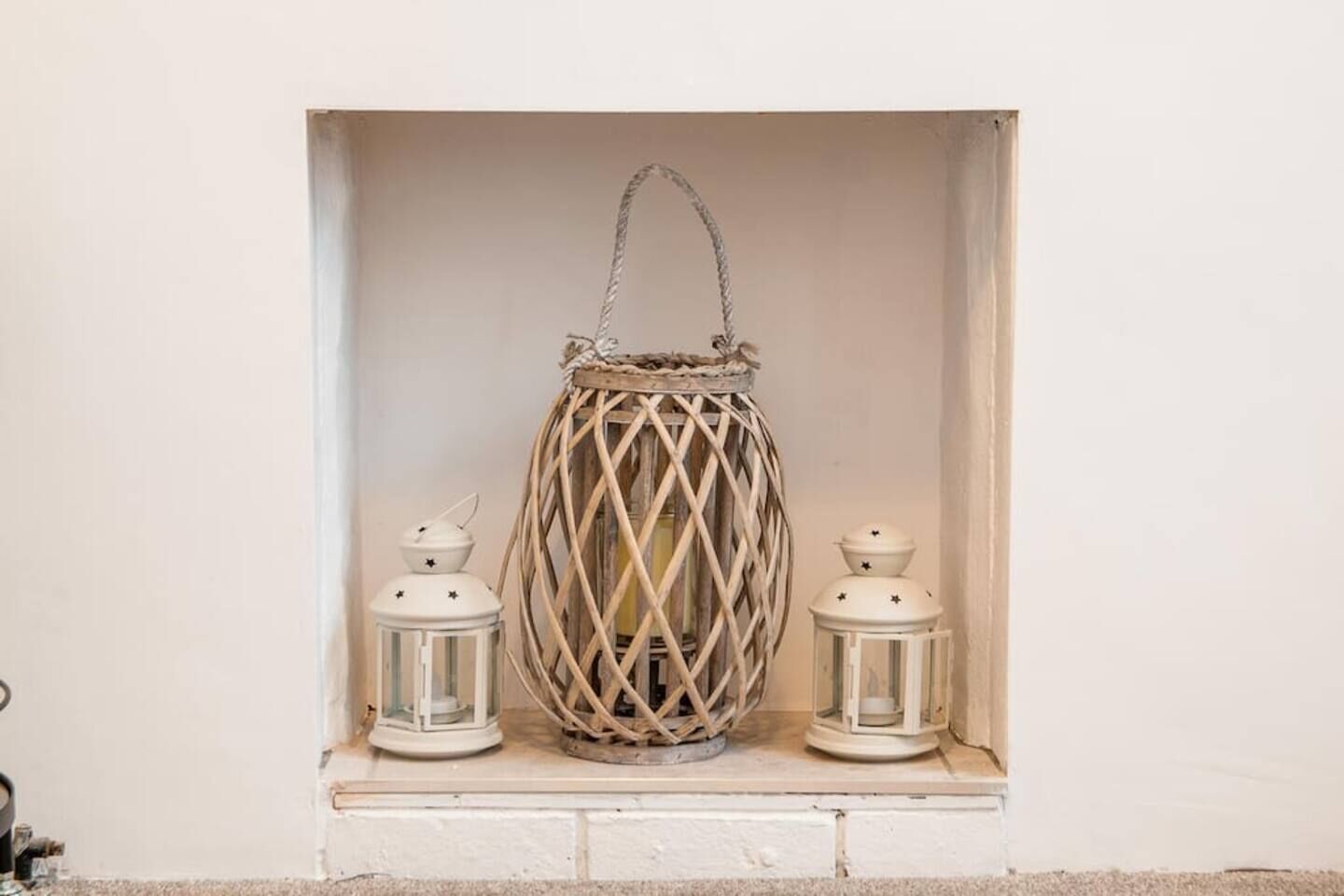 Une lanterne blanche ornée d’un motif tissé, placée dans une alcôve aux murs blancs, et flanquée de deux plus petites lanternes blanches.