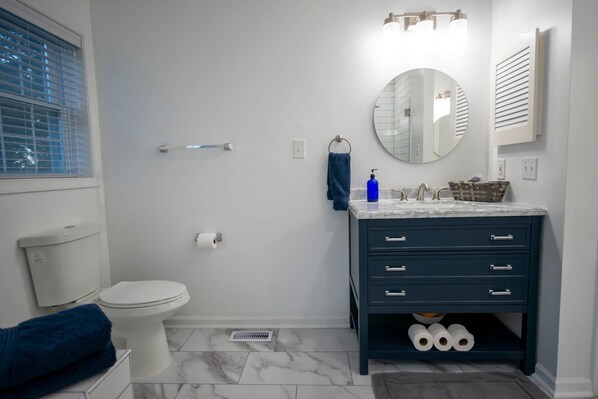 A modern bathroom with a white toilet, a dark blue vanity with drawers, a round mirror, and a towel rack. - Beachfront Getaway, Mermaid Suite, 🐶 💙 (Norfolk)