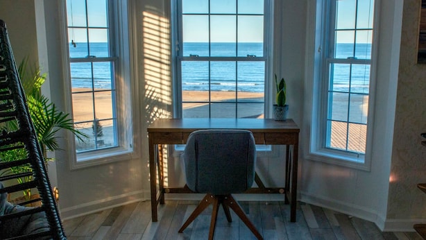 A room with a desk and chair facing large windows overlooking a beach.