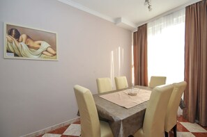 A dining area with a table set for four, beige chairs, a painting on the wall, and a window with curtains.