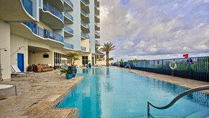 Uma piscina com um elemento aquático azul, área de estar com assentos e um edifício com varandas.