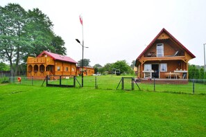 A wooden cabin with a red roof and a smaller adjacent structure, both surrounded by a fenced area and a grassy field. - Wooden House by Ostrowieckie Lake with Beach (Ostrowiec)