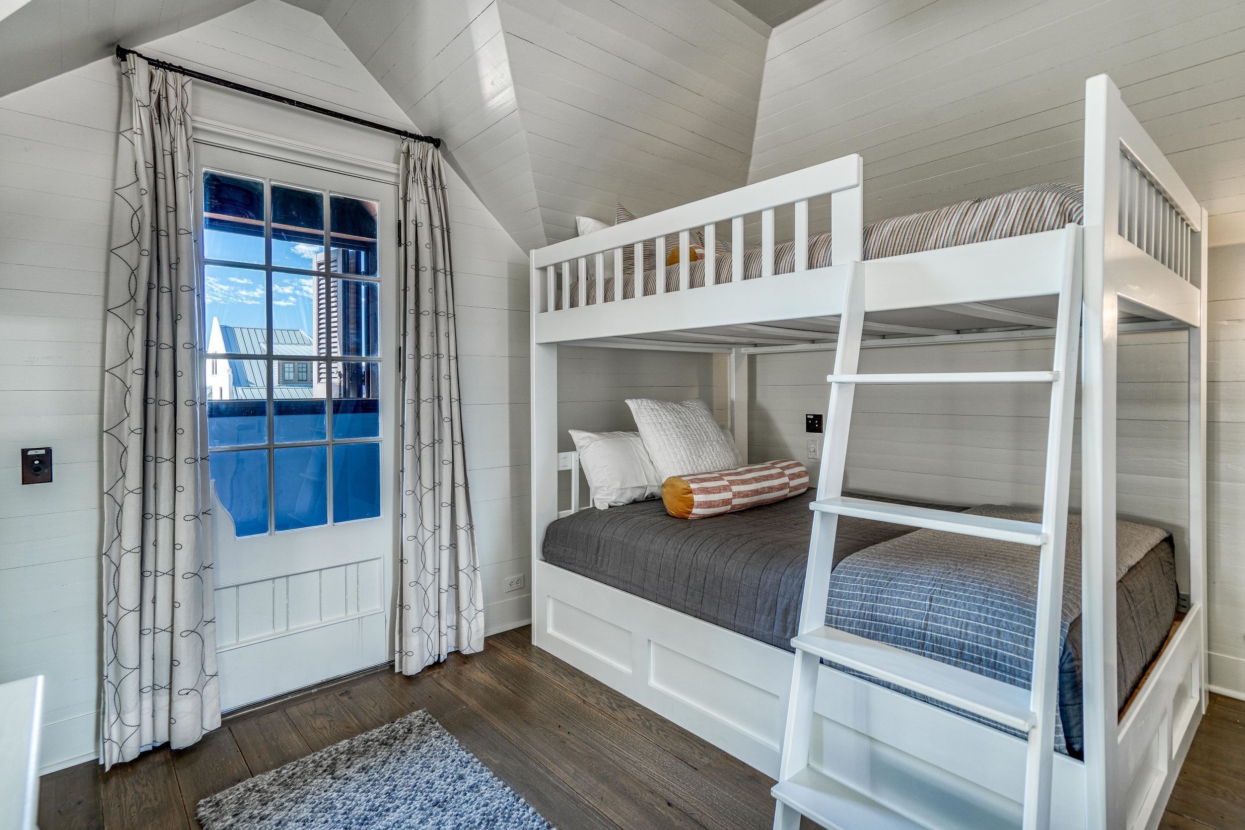 Dormitorio con literas, ventana, cortinas y vista a una casa.