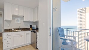 Een keuken met witte kastjes, een vaatwasser en uitzicht op de oceaan vanaf een balkon met een stoel.