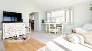 A spacious living room with a flat-screen TV, a wooden coffee table, a white sofa, and a dining area with a table and chairs. - Inviting Oceanfront 3BR Condo in WrightsvilleDunes (Wrightsville Beach)