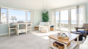 A spacious living room with a large window overlooking the ocean, a white sofa, a wooden coffee table, and a dining table with chairs. - Inviting Oceanfront 3BR Condo in WrightsvilleDunes (Wrightsville Beach)