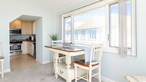 A dining area with a wooden table and chairs, a view of the outside, and a kitchen in the background. - Inviting Oceanfront 3BR Condo in WrightsvilleDunes (Wrightsville Beach)