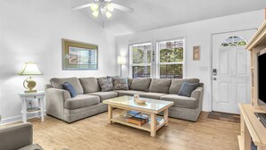 Ein Wohnzimmer mit Ecksofa, Glastisch, Deckenventilator und einer Tür mit Fenster.