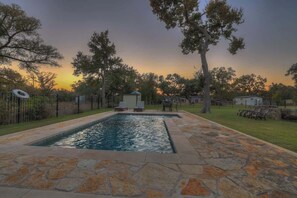 A rectangular swimming pool with stone tiles, surrounded by a stone deck, trees, and a fence. - Dtown Drippin With New Pool! (Dripping Springs)