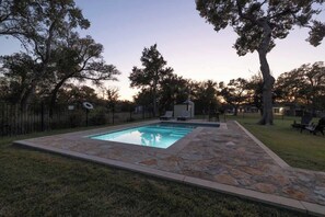 A rectangular swimming pool with stone tiles, surrounded by a grassy area and trees.