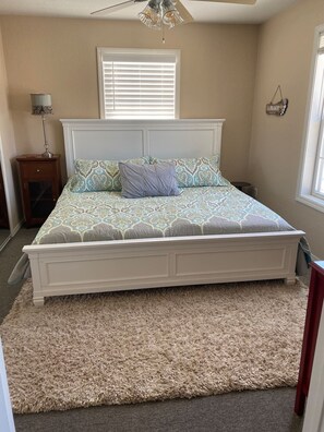 Une chambre avec un lit blanc, une literie à motifs, un tapis beige moelleux, une lampe de chevet et une fenêtre avec des stores.