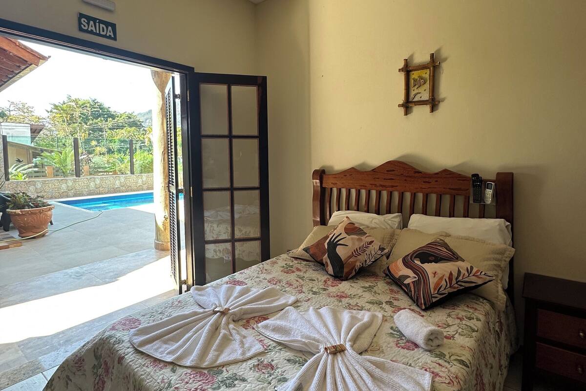 A hotel room with a bed, a wooden headboard, a sign reading "SAÍDA", and a view of a swimming pool outside.