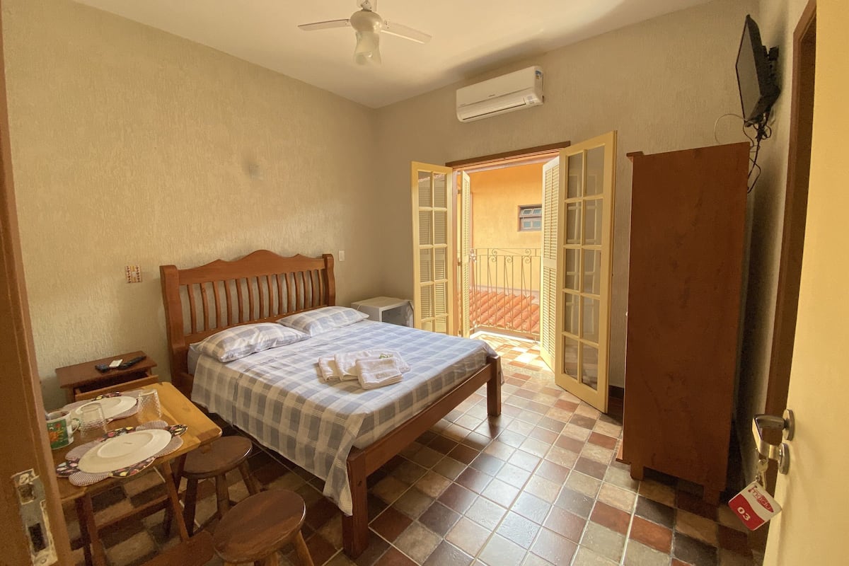 A bedroom with a bed, a wooden wardrobe, a desk with a chair, and a balcony with a view.