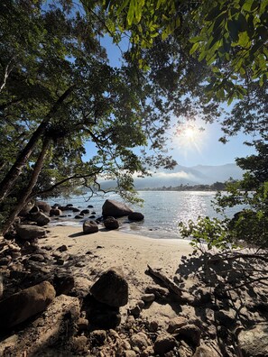 A beach with rocks and trees, a clear sky, and a distant mountain range. - Pousada Madu Eireli (Ubatuba)