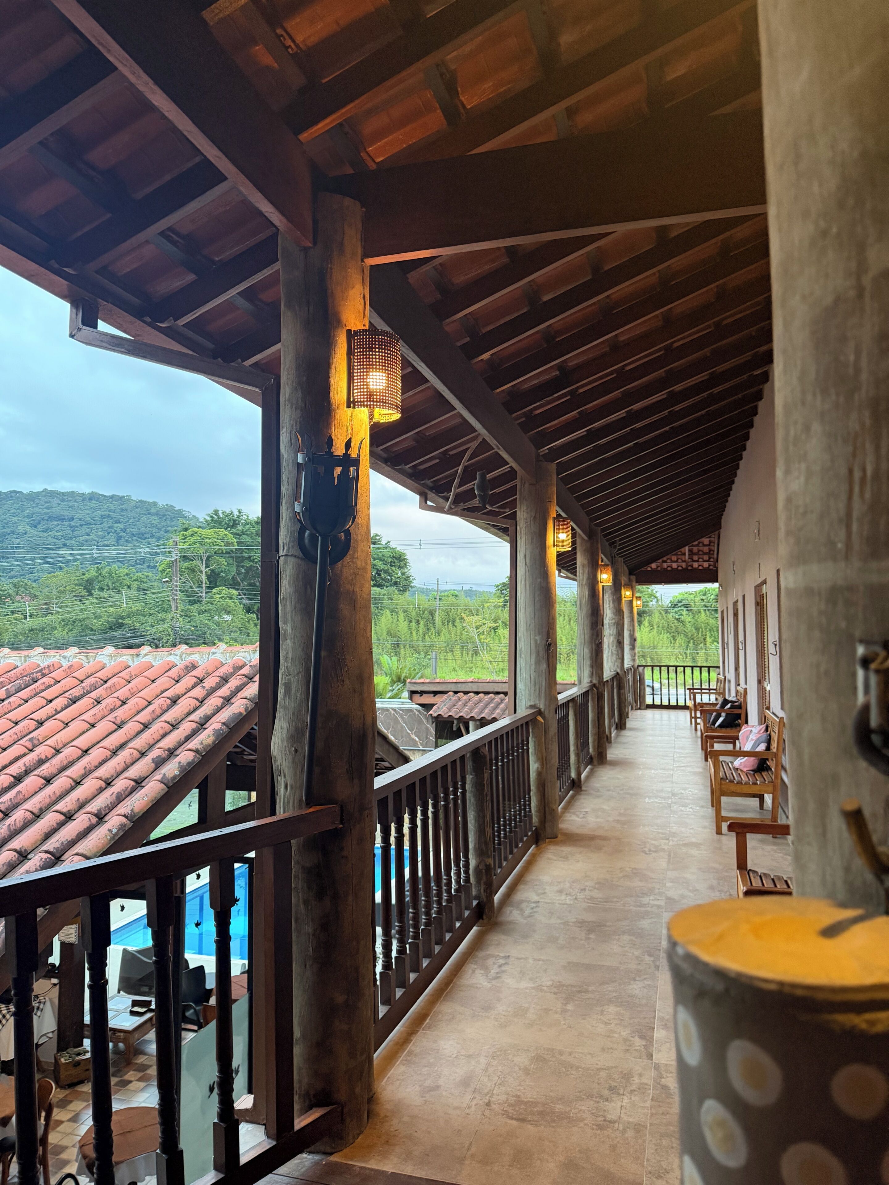 A covered outdoor area with wooden beams and a tiled roof, benches, and a view of a hilly landscape.