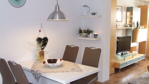 A modern dining area with a white table, brown chairs, and a pendant light. There is a kitchen visible in the background with a TV and cabinets. - Hochwald vacation apartment - in the heart of the Saarland (Quierschied)