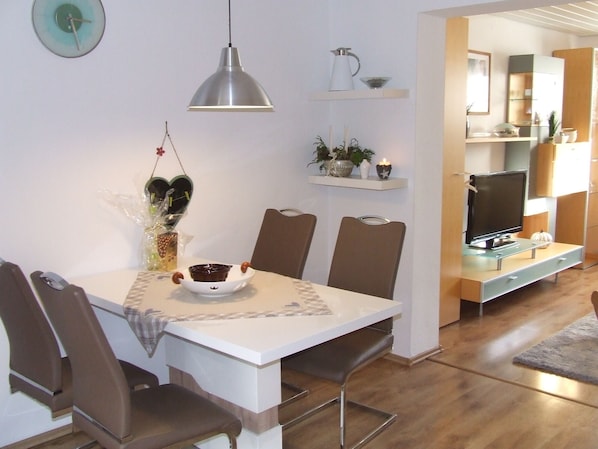 A modern dining area with a white table, brown chairs, and a pendant light. There is a kitchen visible in the background with a TV and cabinets.