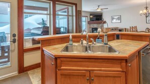 A kitchen with wooden cabinets, a double sink, and a view of the outside through large windows. - Mountain Time-Spacious, Private Hot Tub,Great View (Big White)