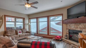 A cozy living room with a fireplace, large windows, a ceiling fan, and a flat-screen TV. - Mountain Time-Spacious, Private Hot Tub,Great View (Big White)