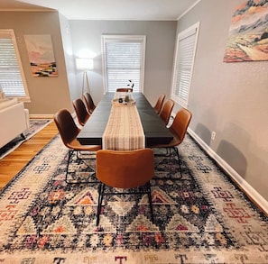 Une salle à manger avec une longue table, six chaises, un tapis à motifs et deux fenêtres équipées de stores.