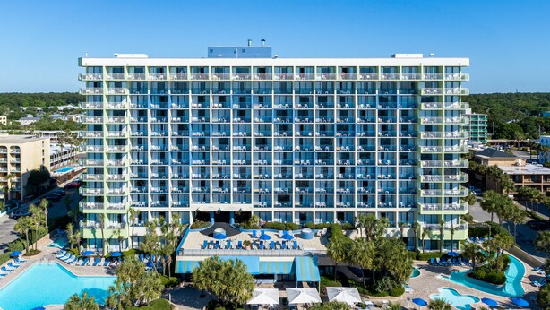 A large hotel with multiple balconies, a swimming pool, and a landscaped area with palm trees. - Family Suite at Coral Beach Resort - Full Access to Amenities! (Myrtle Beach)