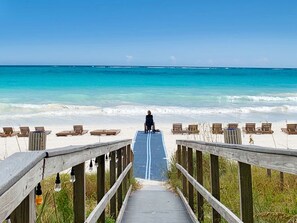 Ein Holzsteg führt zu einem Strand mit Liegestühlen und einer Person im Rollstuhl.