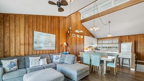 A cozy living room with a sectional sofa, a dining area with light blue chairs, and a kitchen with wooden walls and white cabinets.
