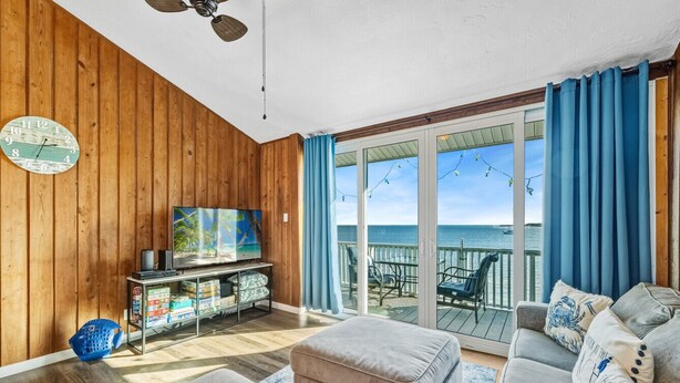 A living room with a wooden wall, a television, a sofa, and a view of the ocean through large glass doors.