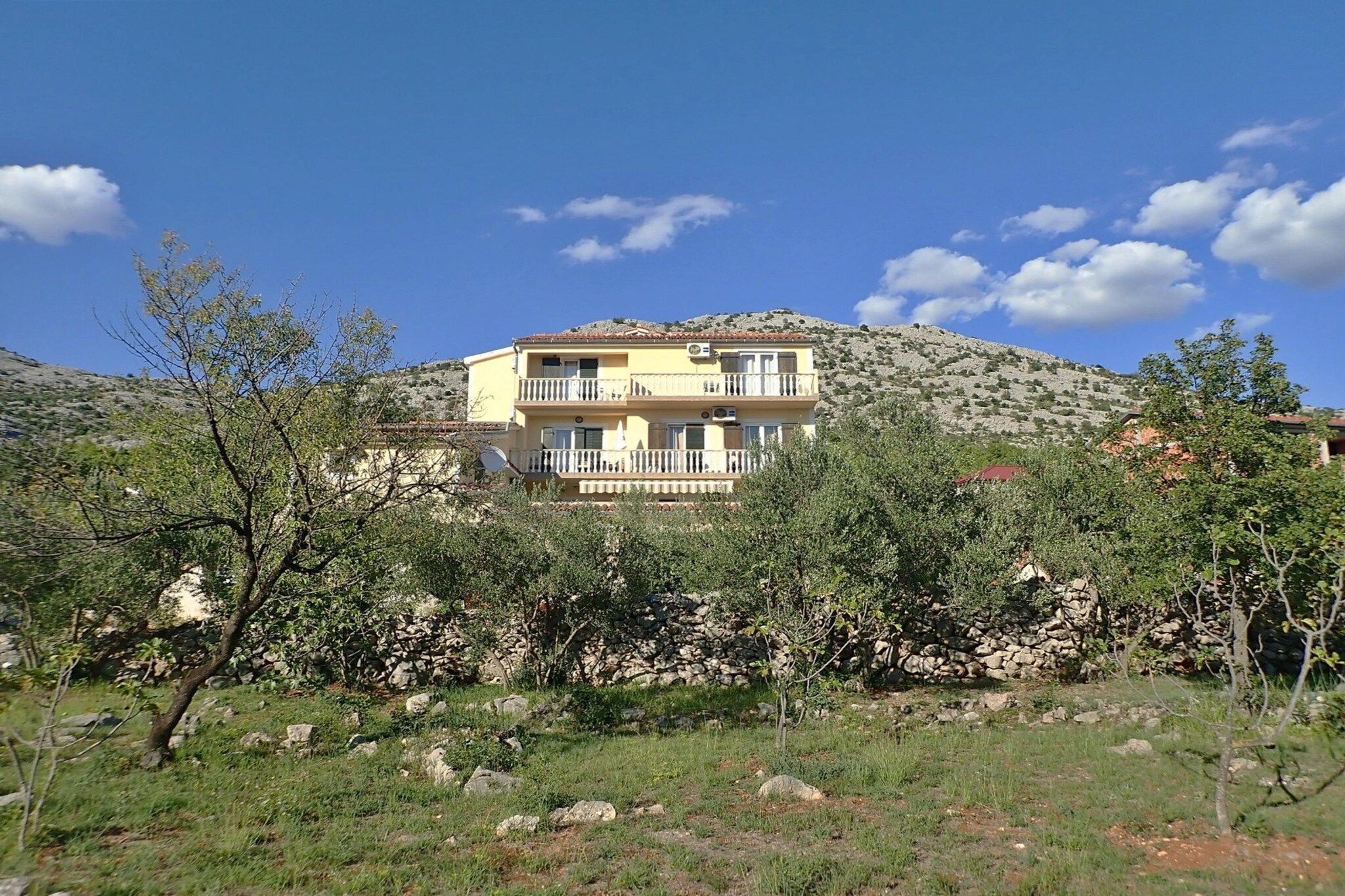 A multi-story building with balconies, surrounded by trees and a stone wall.