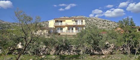 A multi-story building with balconies, surrounded by trees and a stone wall.