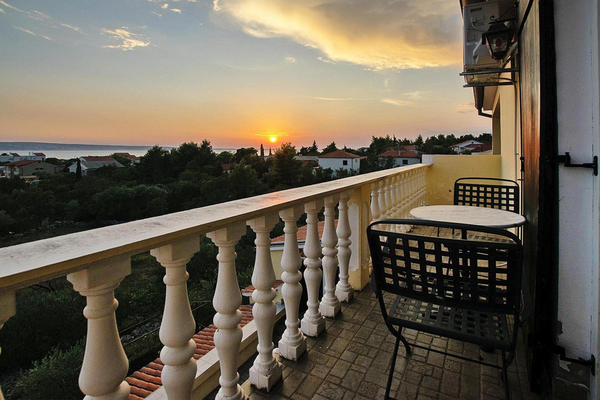 A balcony with a railing, a table, and a chair, overlooking a residential area and the sea during sunset.