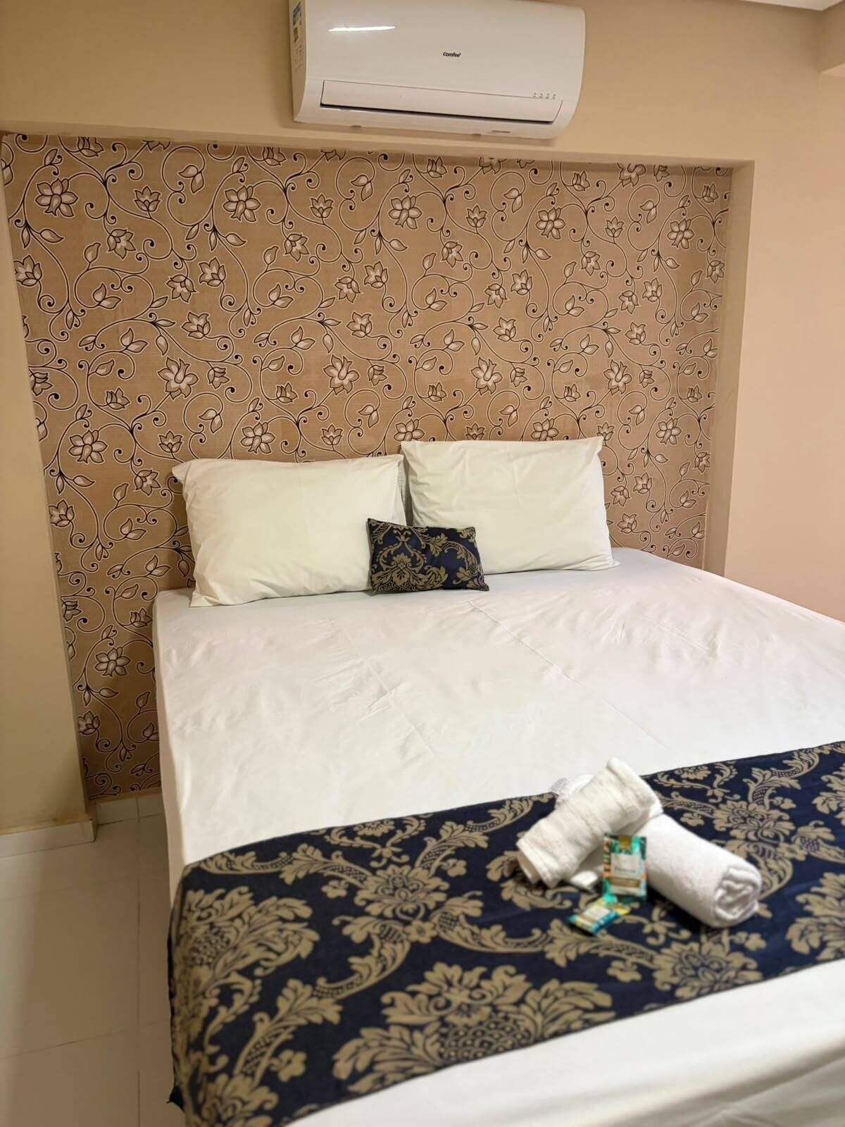 A hotel room with a bed, a decorative headboard, a wall-mounted air conditioner, and a neatly made bed with a patterned bedspread and pillows.