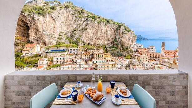 A balcony with a table set for breakfast overlooking a coastal town with a prominent cliff. - Maika House - Amalfi Coast - seaview (Atrani)