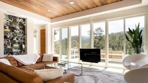 A modern living room with a large window, a flat-screen TV, and a brown leather sofa. - Modern Luxury - Carmel (Carmel-by-the-Sea)