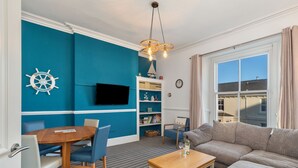 Uma sala de estar com parede azul, uma mesa de jantar de madeira com cadeiras, um sofá, uma mesa de centro, uma estante de livros e uma TV fixada na parede.